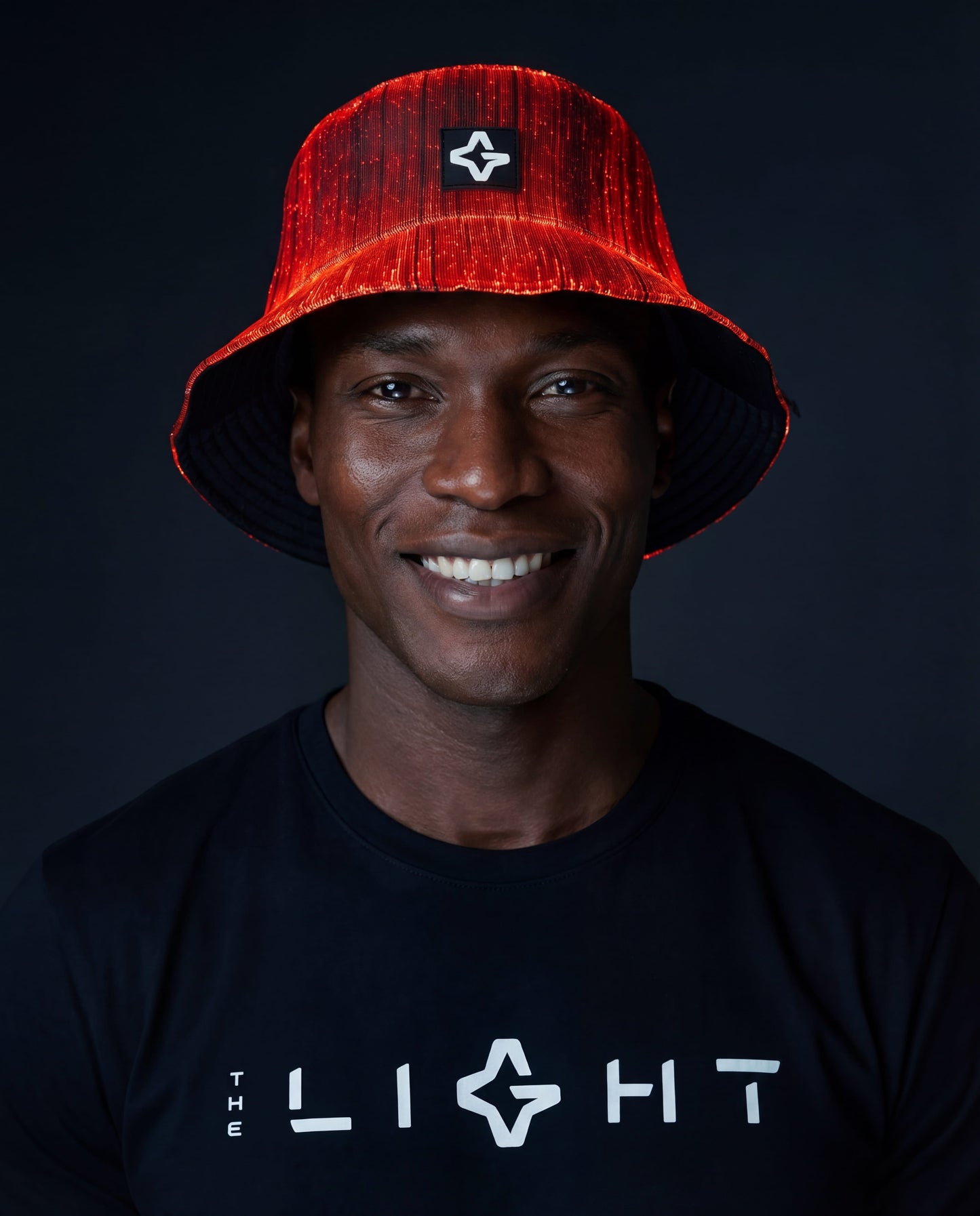Man wearing a red illuminated fiber-optic bucket hat and a black T-shirt with “THE LIGHT” logo against a dark background