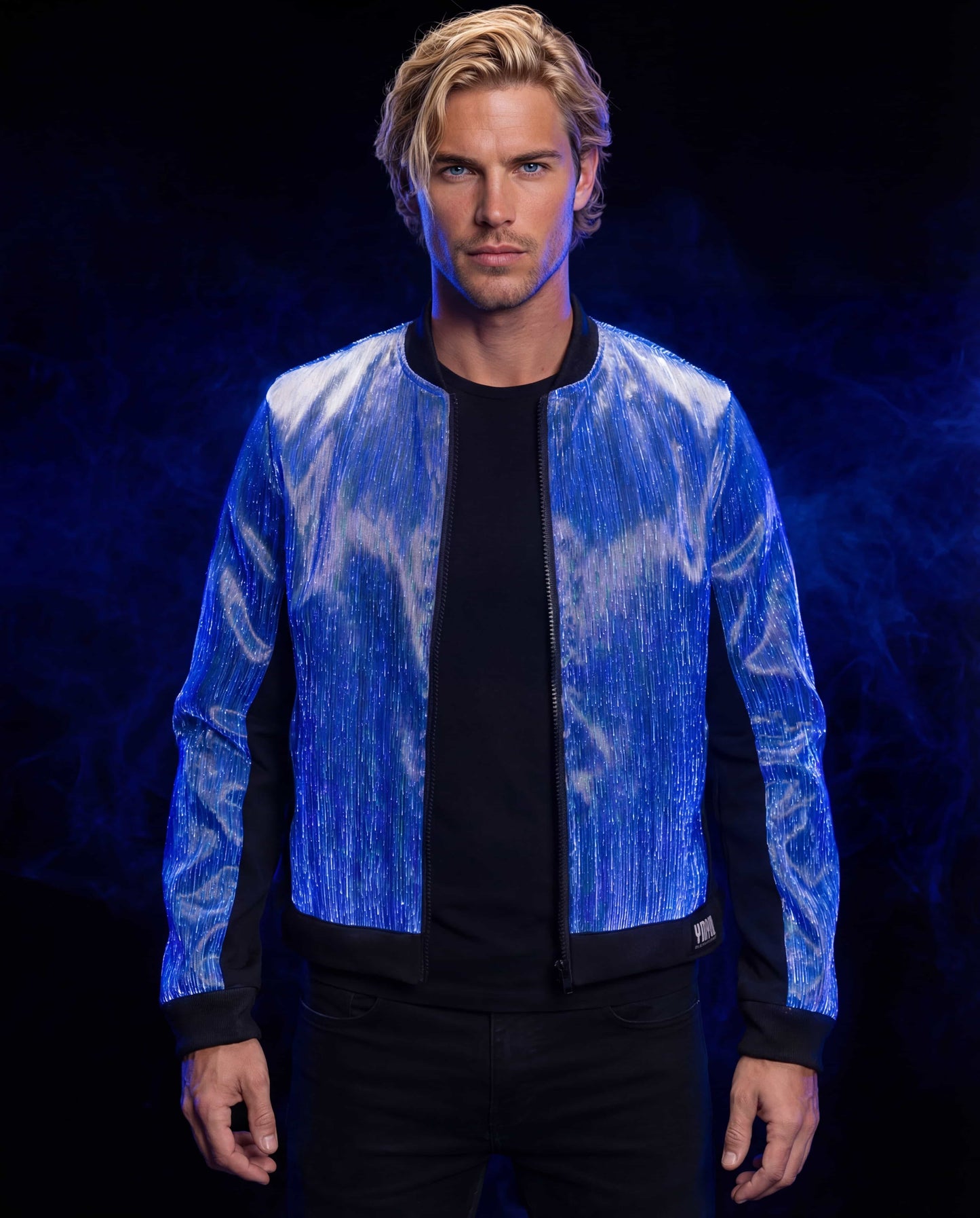 Man wearing a glowing blue fiber-optic bomber jacket, standing against a dark blue smoke-filled studio background