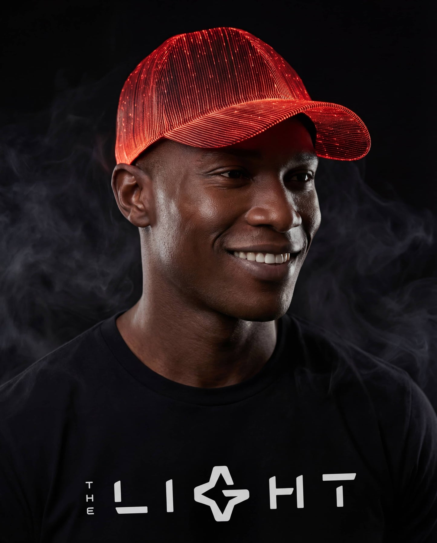 Man wearing a glowing red fiber-optic snapback hat, smiling and standing against a dark smoky background while wearing a black THE LIGHT shirt.