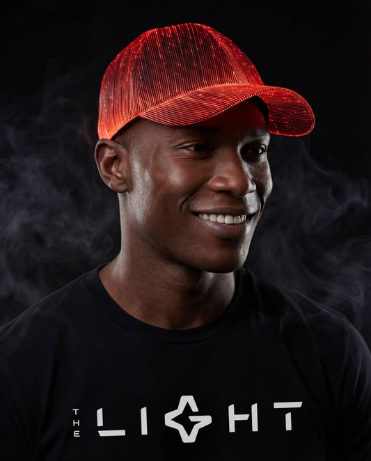 Man wearing a glowing red fiber-optic snapback hat, smiling and standing against a dark smoky background while wearing a black THE LIGHT shirt.