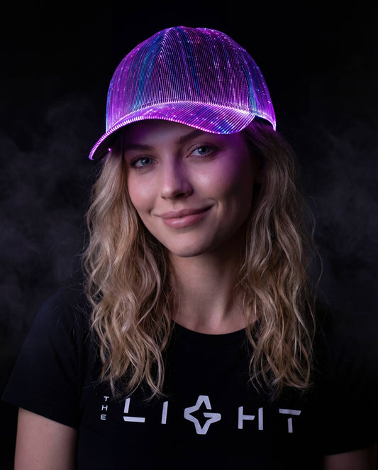 Woman wearing a glowing multicolor fiber-optic LED snapback hat, smiling against a dark smoky background while wearing a black THE LIGHT shirt