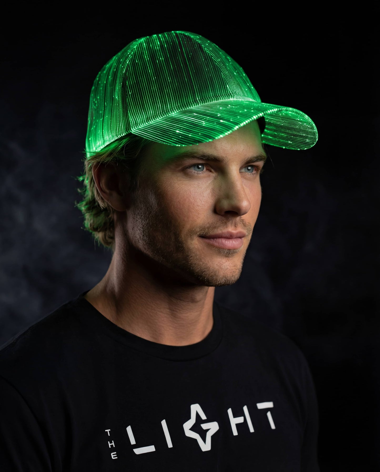 Man wearing a glowing green fiber-optic LED snapback hat, looking to the side against a dark smoky background while wearing a black THE LIGHT shirt