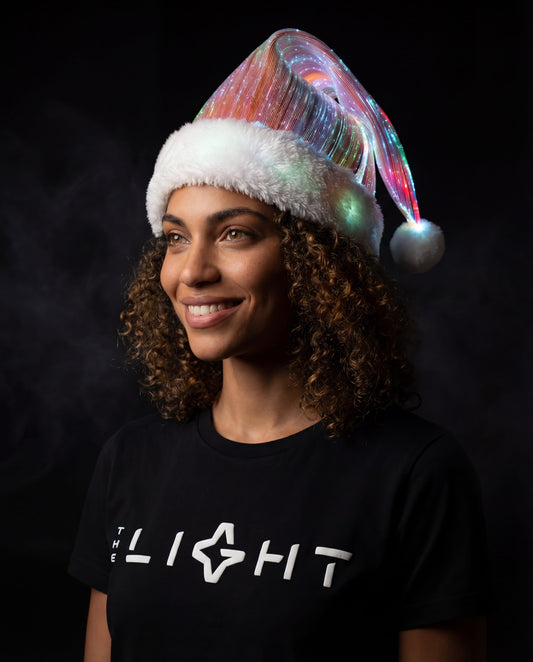Person wearing a fiber optic Christmas hat with multicolor illumination, smiling while wearing a black THE LIGHT shirt against a dark background