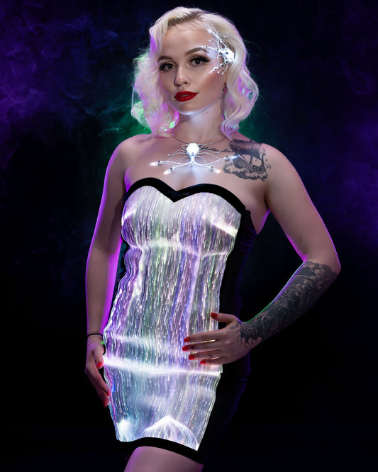 Woman wearing a white fiber optic light up dress glowing in multicolor light against a dark background