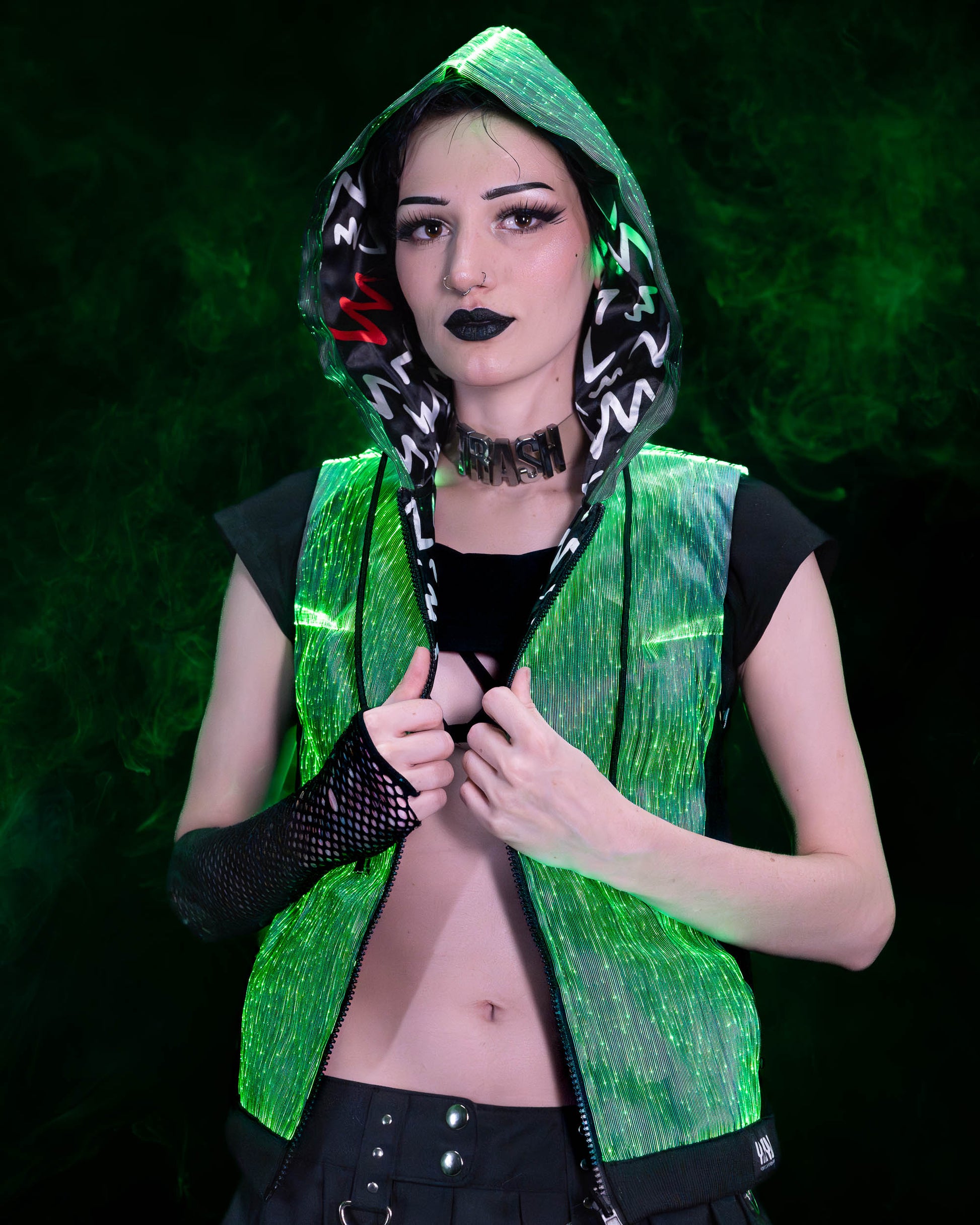 Woman wearing a black fiber optic light up sleeveless hoodie glowing in green light against a dark background
