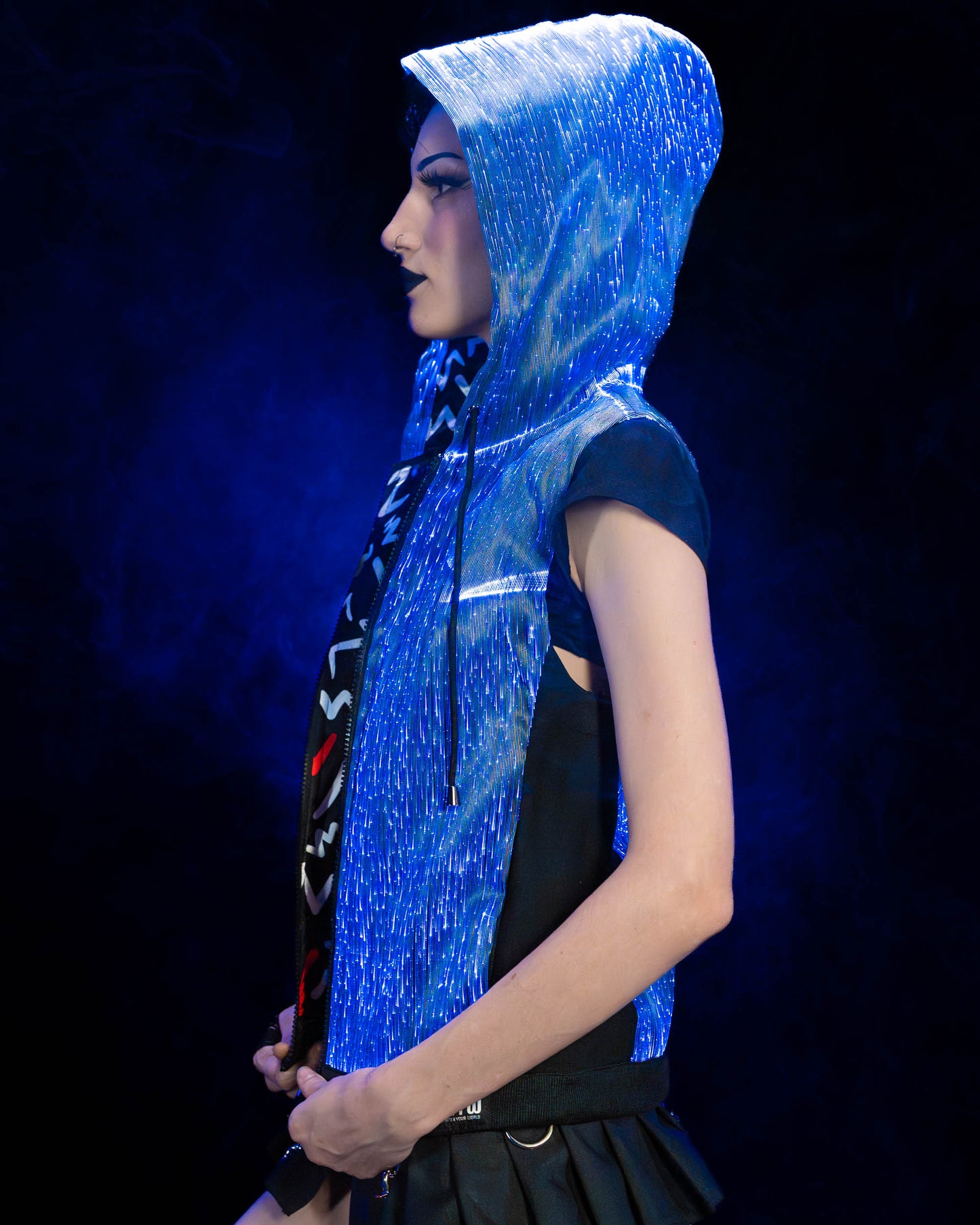 Woman wearing a black fiber optic light up sleeveless hoodie glowing in blue light  from the side against a dark background