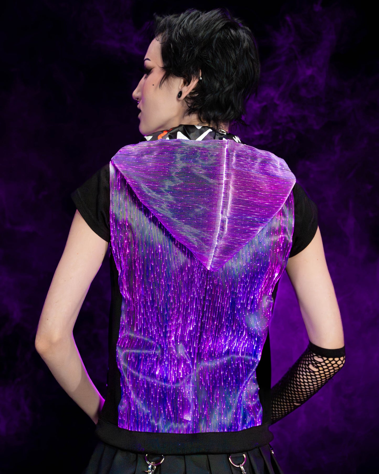 Woman wearing a black fiber optic light up sleeveless hoodie glowing in purple light seen from the back against a dark background