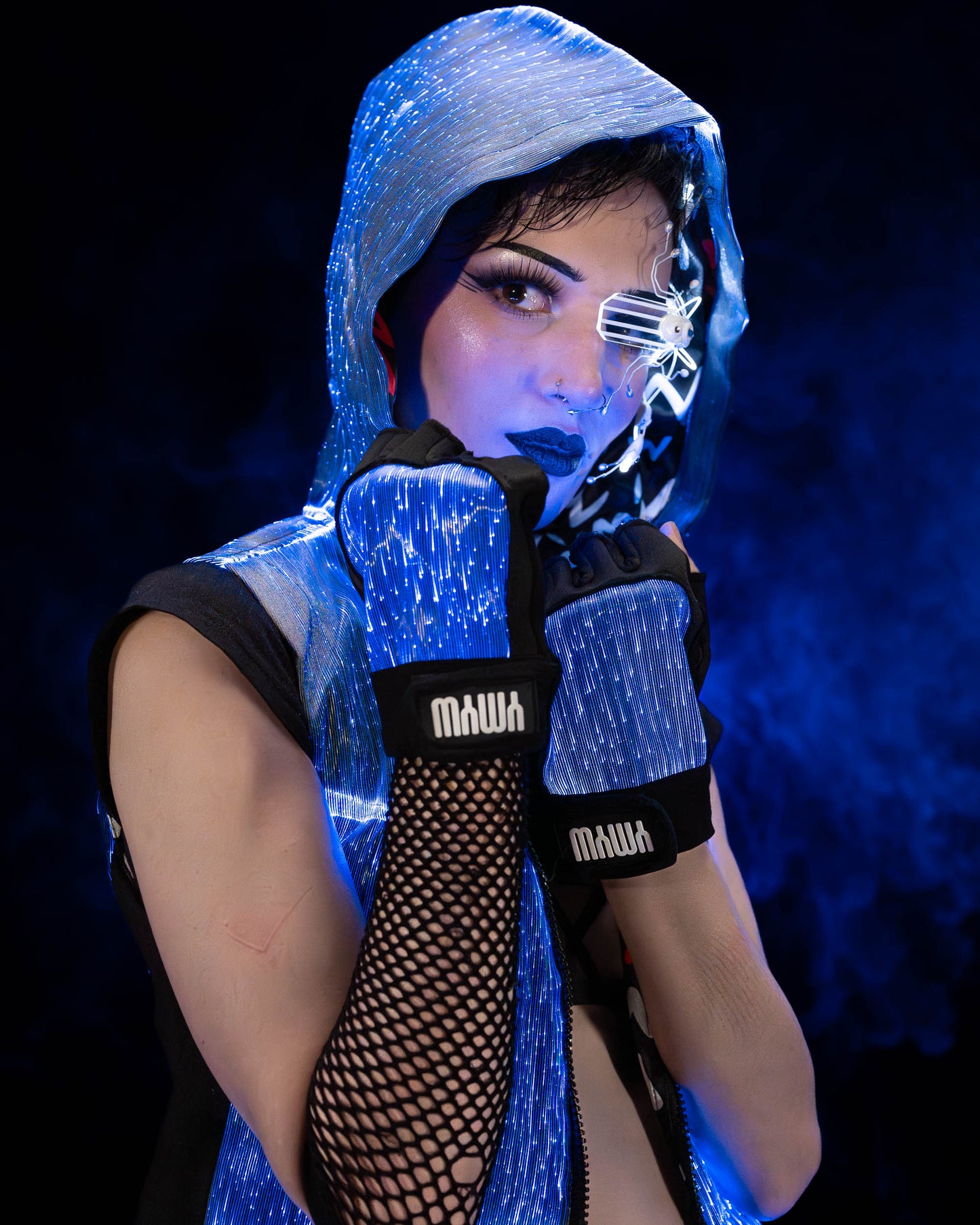Woman wearing a black fiber optic light up sleeveless hoodie and gloves illuminated in blue light against a dark background