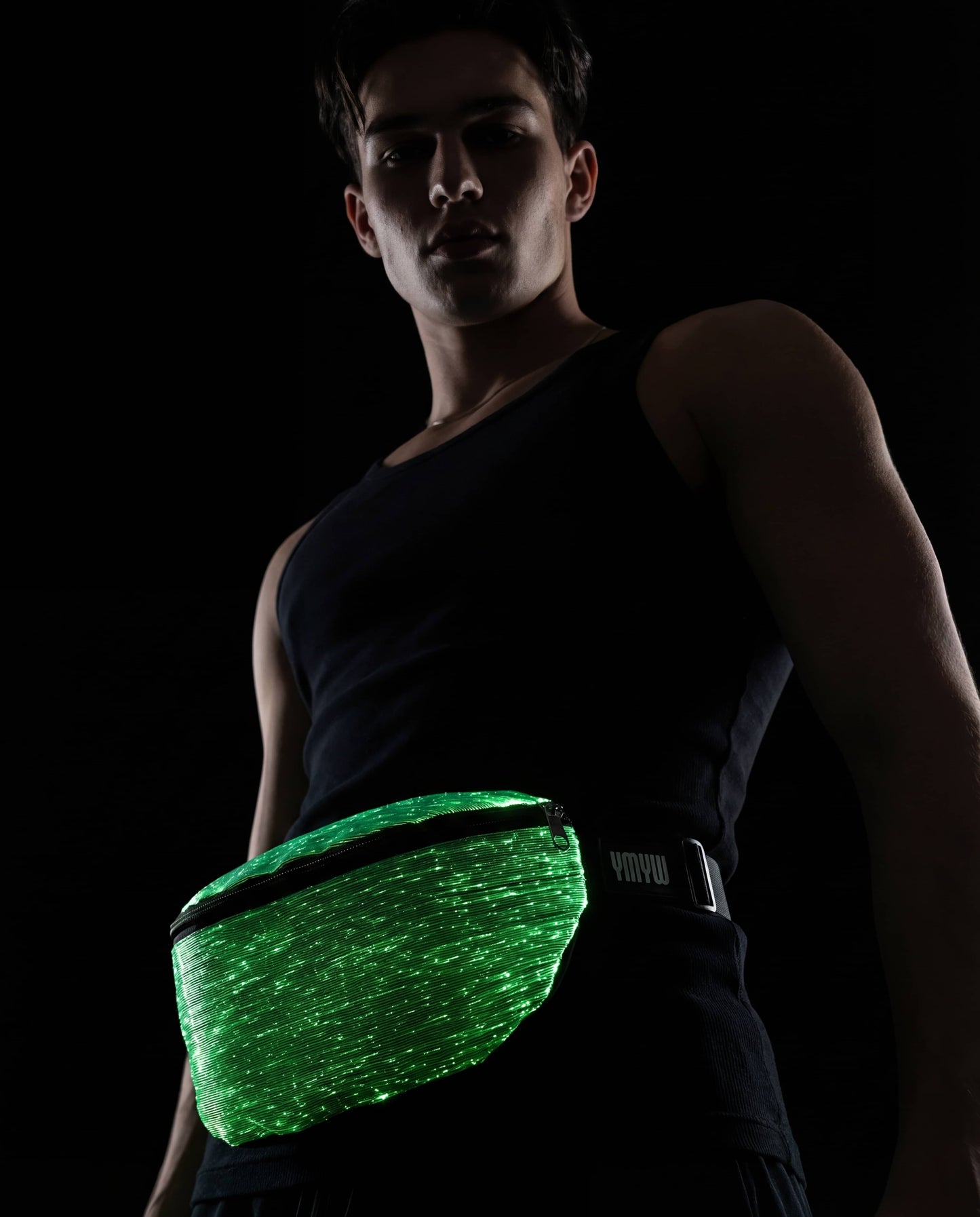 Man wearing a black tank top with a glowing green fiber-optic fanny pack, posed against a dark background