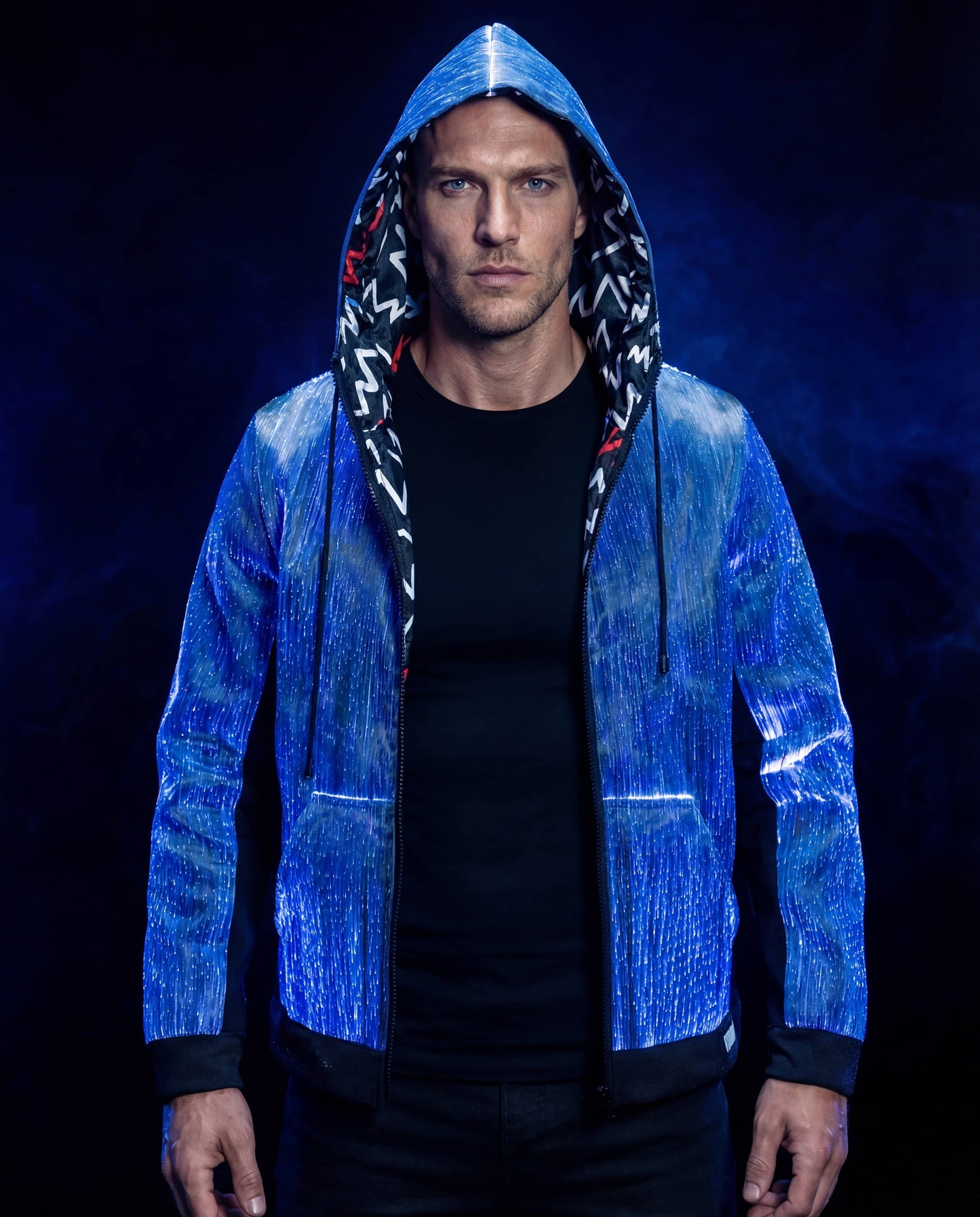 Man wearing a glowing blue fiber-optic hoodie with the hood up, standing against a dark blue smoke-filled background