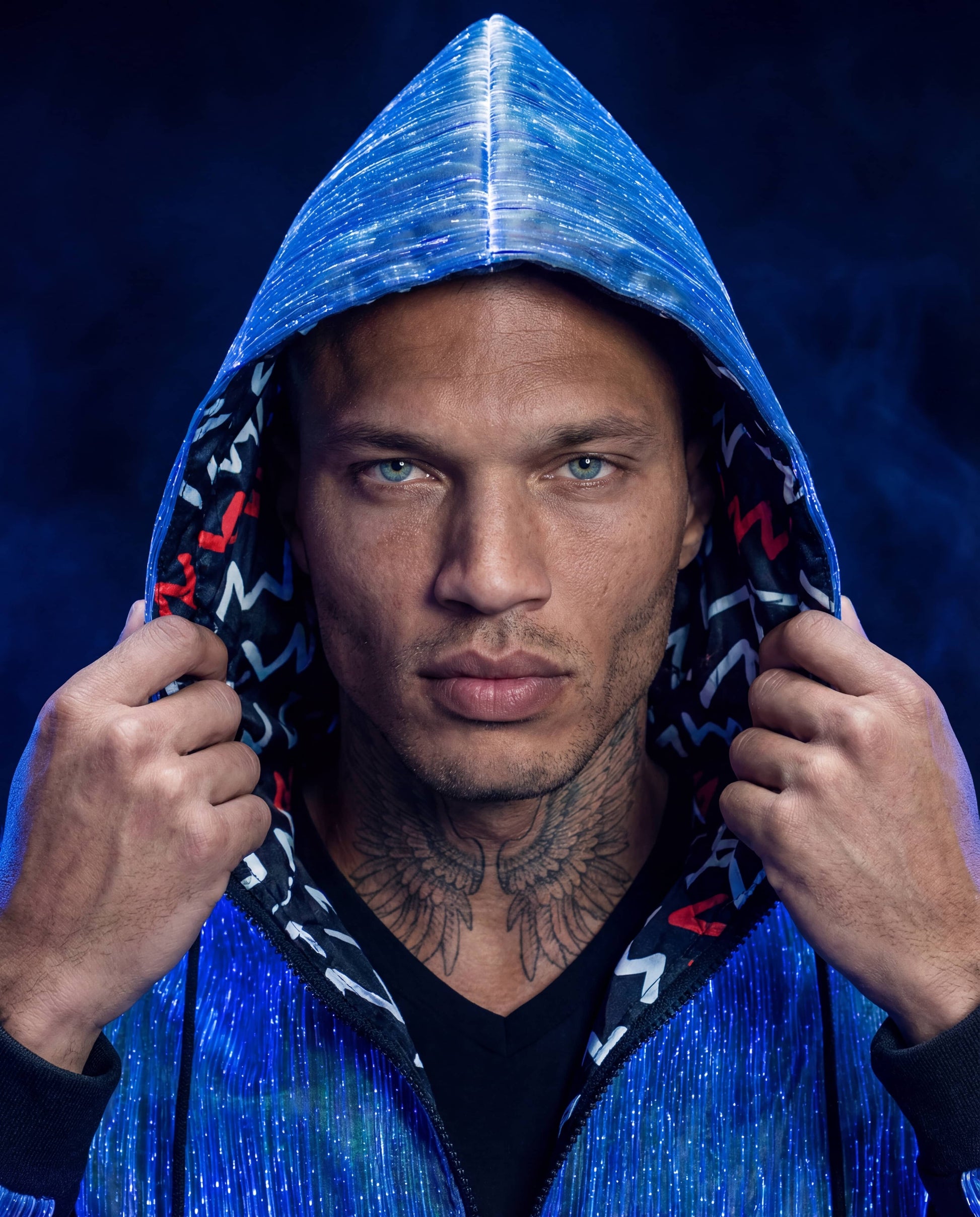 Man wearing a glowing blue fiber-optic hoodie, holding the hood with both hands against a dark blue smoke-filled background.
