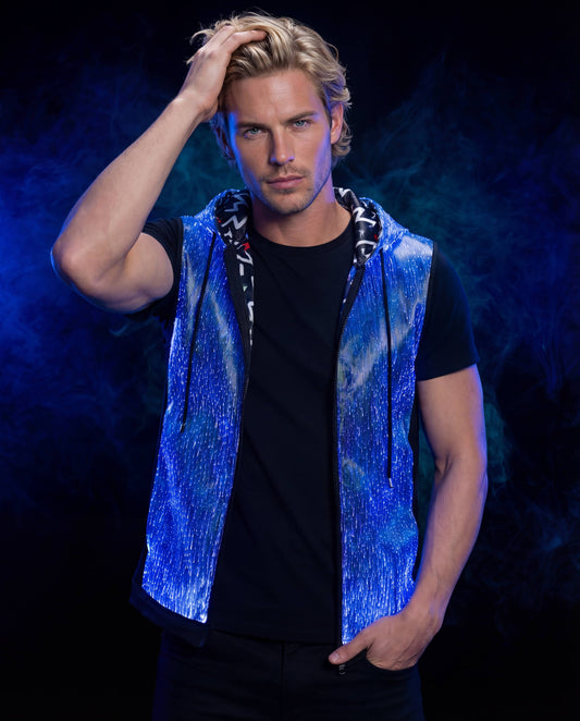 Man wearing a glowing blue sleeveless fiber-optic hoodie, standing against a dark blue smoke-filled background with one hand in his hair