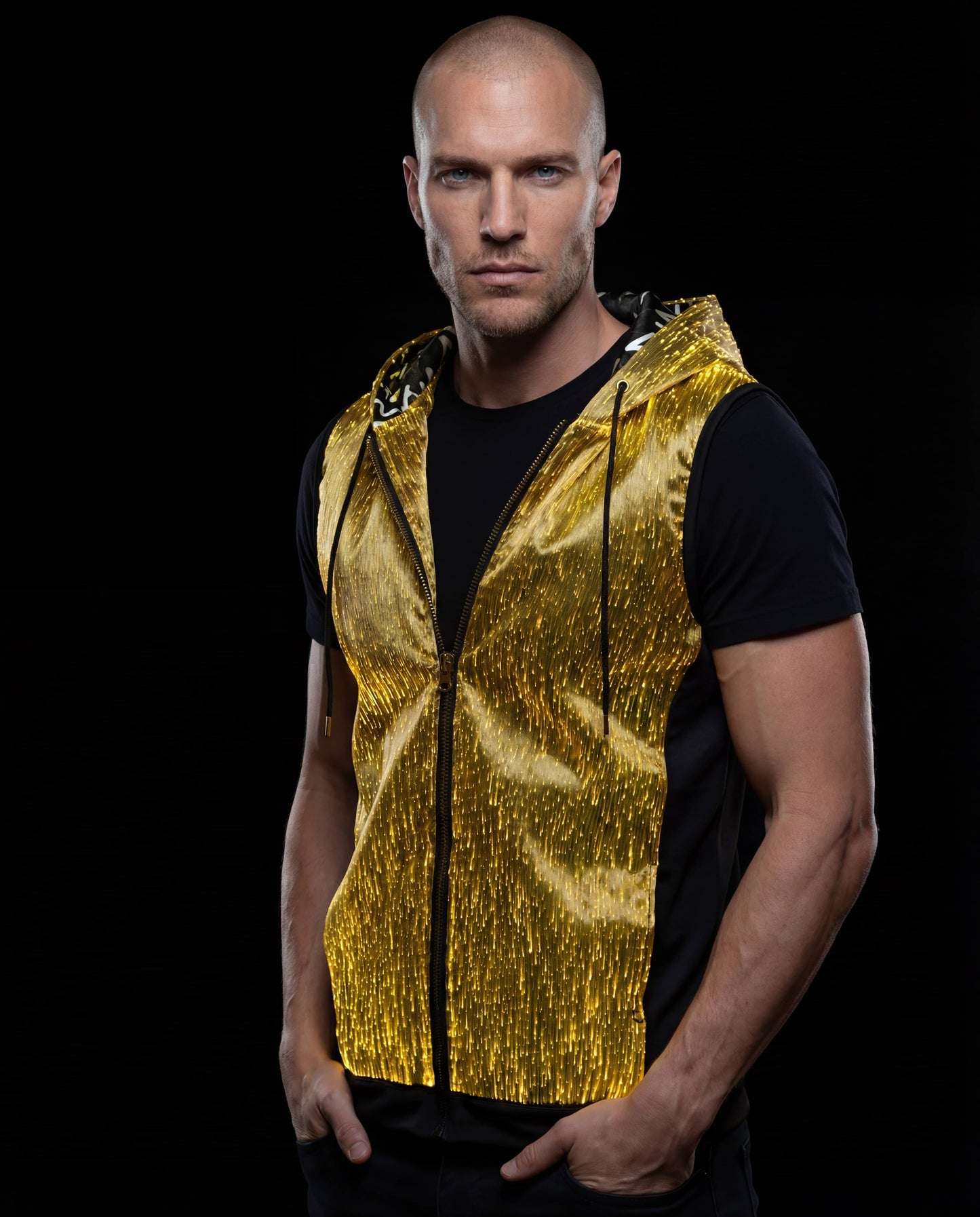 Man wearing a glowing gold sleeveless fiber-optic hoodie, standing against a black background with his hands in his pockets
