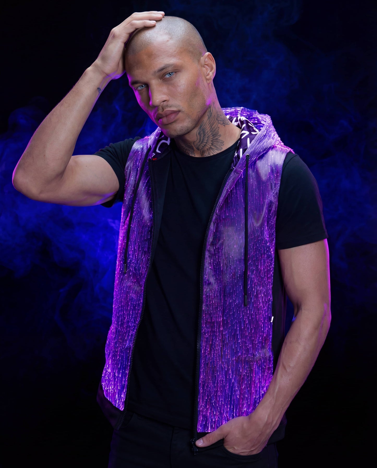 Man wearing a glowing purple sleeveless fiber-optic hoodie, standing against a dark blue smoke-filled background with one hand on his head