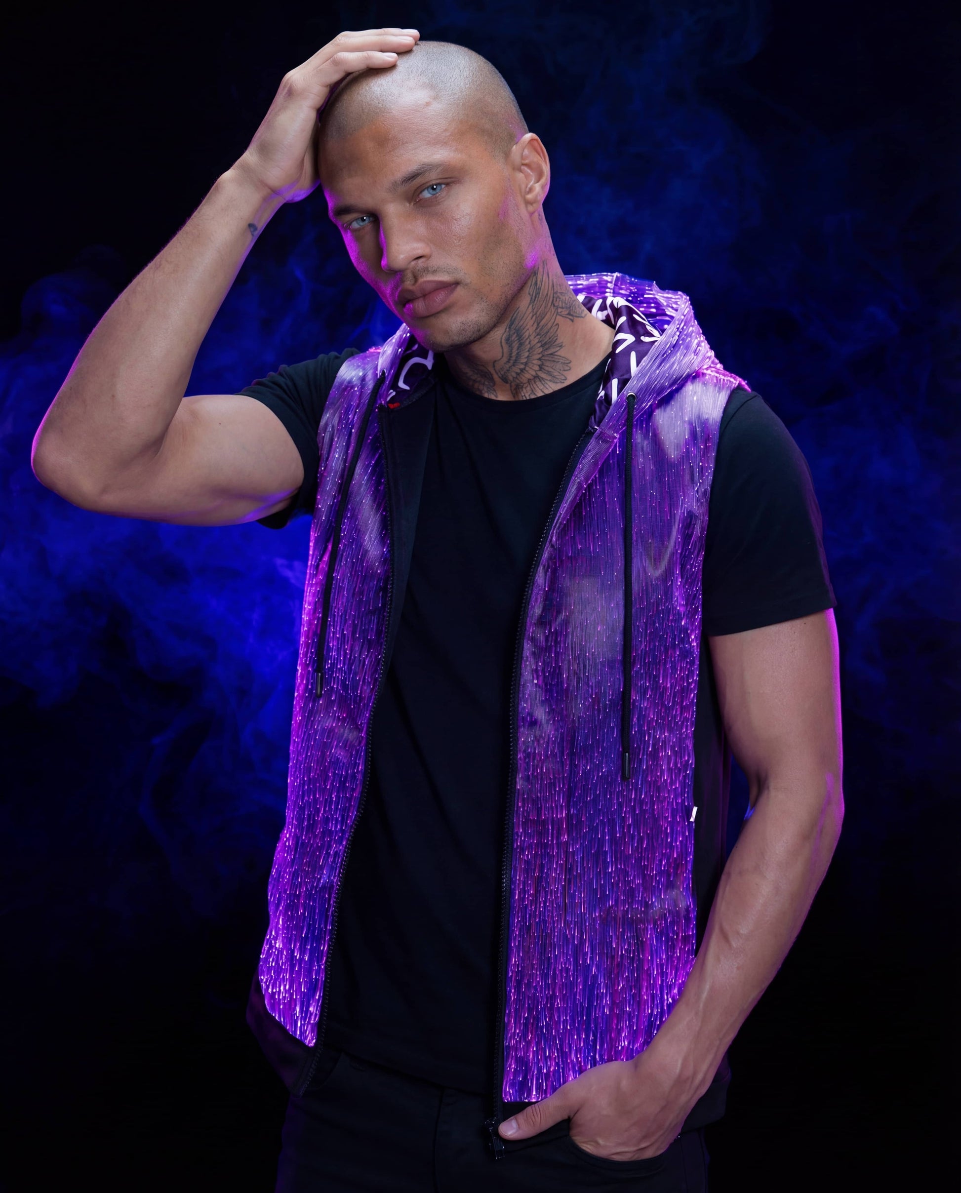 Man wearing a glowing purple sleeveless fiber-optic hoodie, standing against a dark blue smoke-filled background with one hand on his head