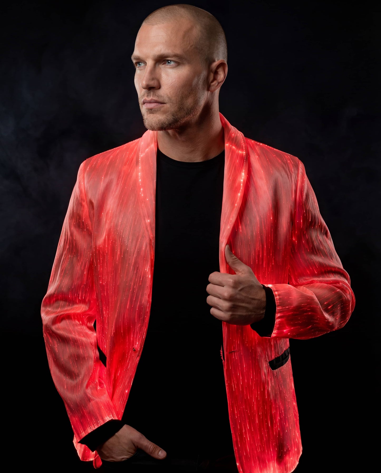 Man wearing a glowing red fiber-optic suit jacket, standing against a dark smoky background with one hand adjusting the lapel