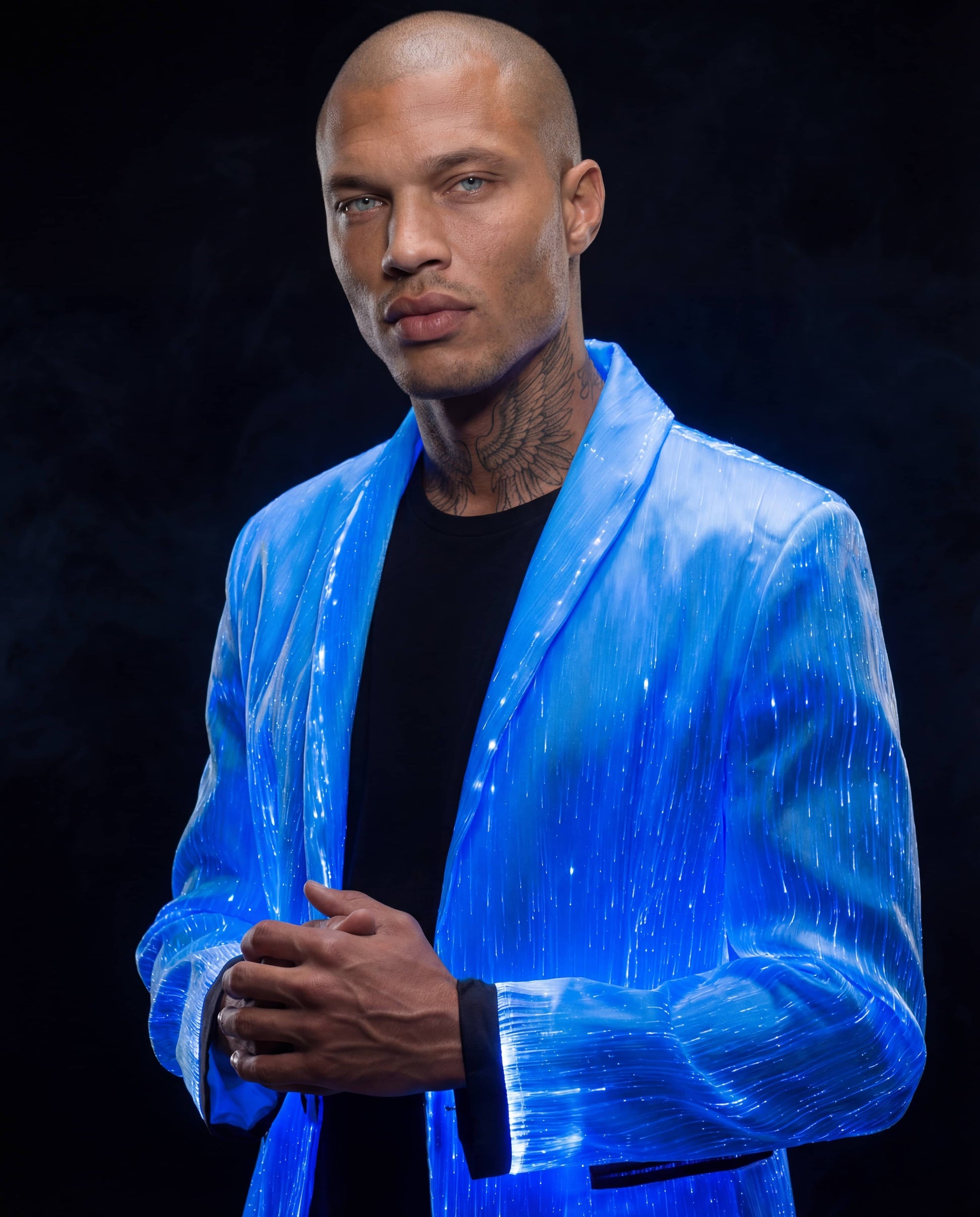 Man wearing a glowing blue fiber-optic suit jacket, standing against a dark smoky background with his hands lightly clasped in front