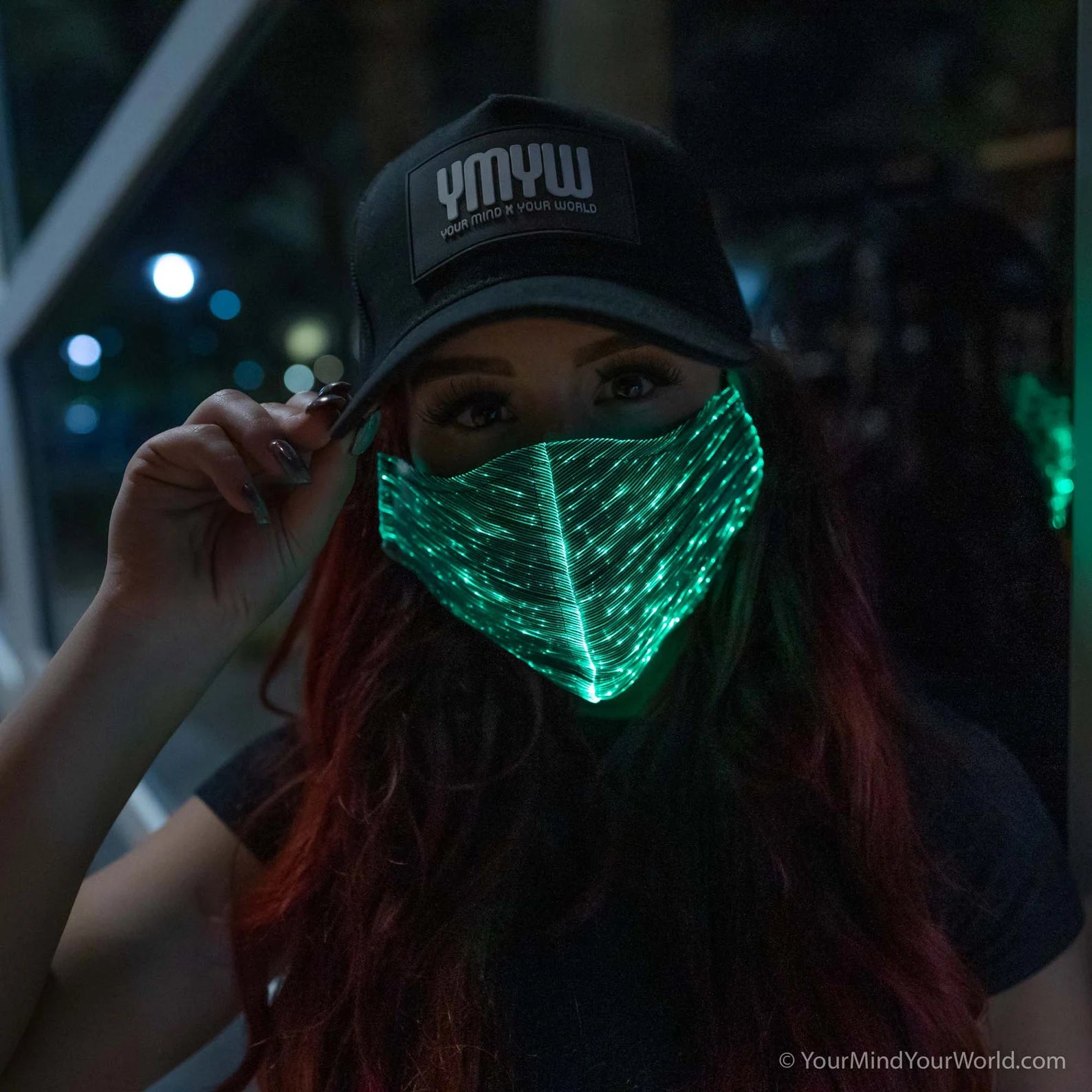 Woman wearing a YMYW cap and a fiber optic light-up mask illuminated in green light against a dark background