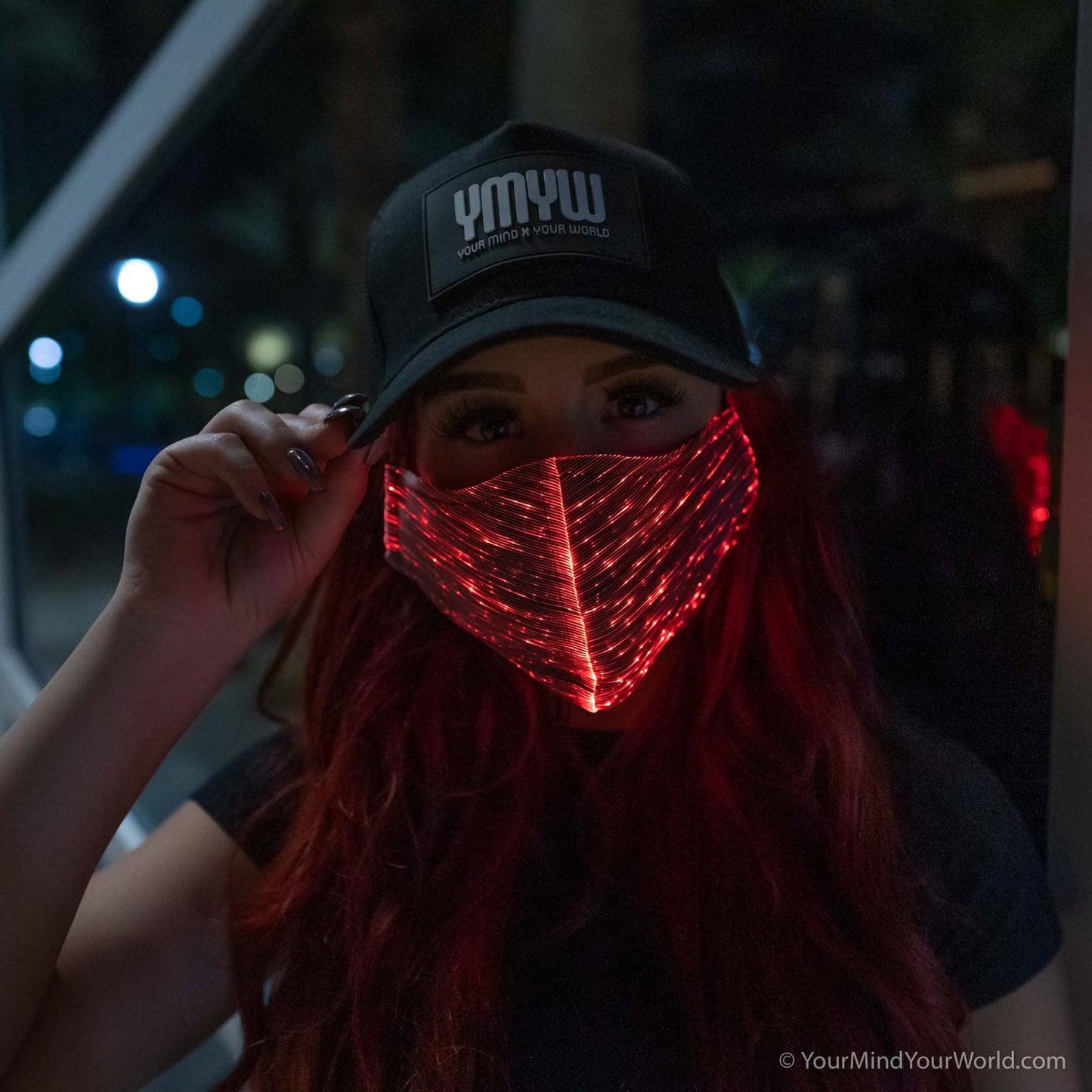 Woman wearing a YMYW cap and a fiber optic light-up mask illuminated in red light against a dark background
