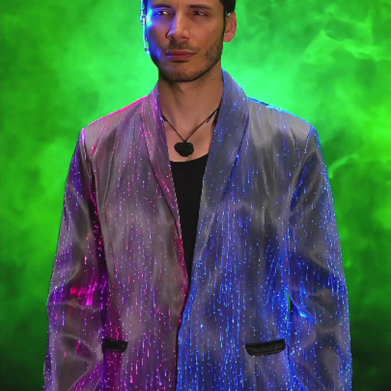 Man wearing a black fiber optic LED suit jacket illuminated in shifting colors, standing against a smoky gradient background. The jacket glows through embedded optic fibers with changing through colors such as blue, pink, and purple light.
