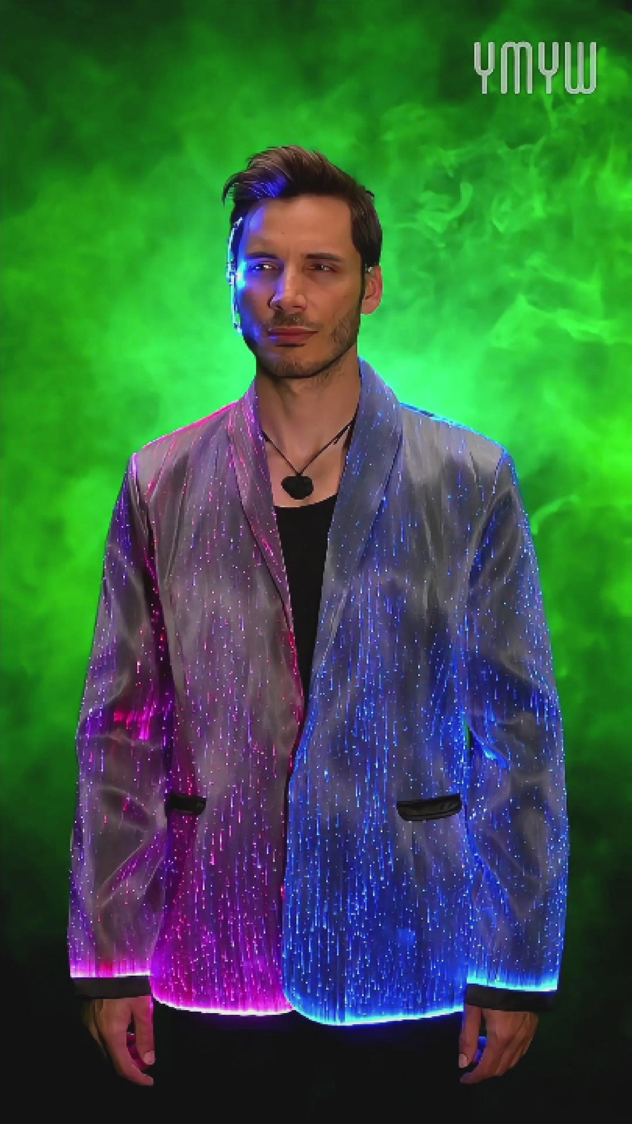 Man wearing a black fiber optic LED suit jacket illuminated in shifting colors, standing against a smoky gradient background. The jacket glows through embedded optic fibers with changing through colors such as blue, pink, and purple light.