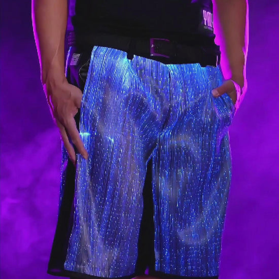 Man wearing black fiber-optic LED light-up shorts rotating on a platform, the fabric glowing in shifting colors against a smoky background