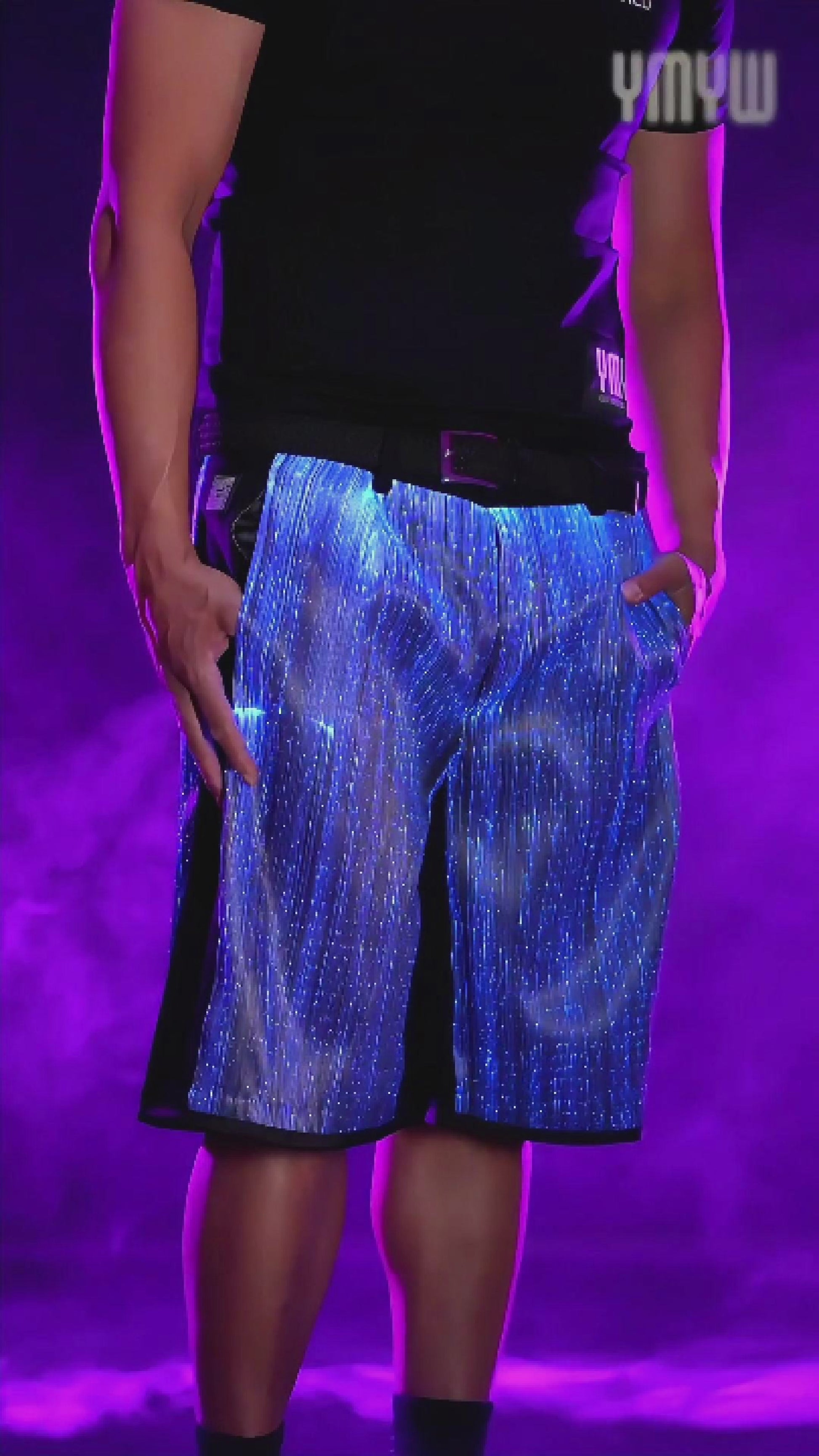 Man wearing black fiber-optic LED light-up shorts rotating on a platform, the fabric glowing in shifting colors against a smoky background