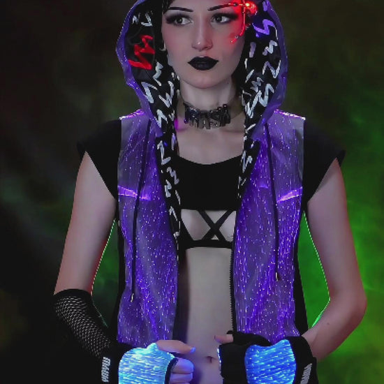 Woman wearing a black sleeveless fiber-optic LED hoodie glowing in various colors, standing against a dark colorful backdrop