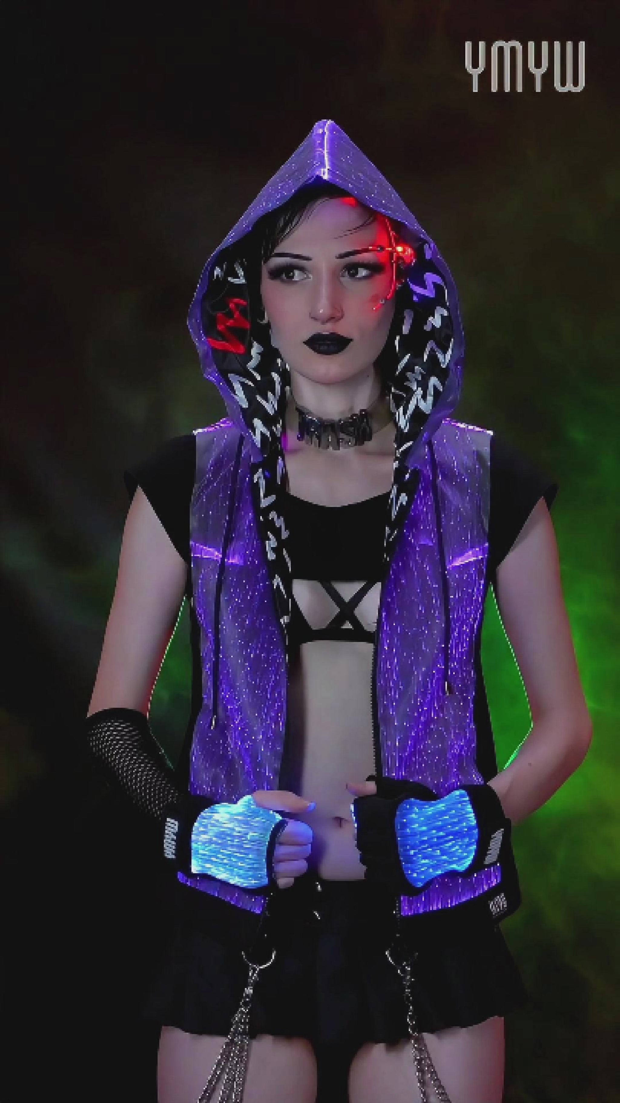 Woman wearing a black sleeveless fiber-optic LED hoodie glowing in various colors, standing against a dark colorful backdrop